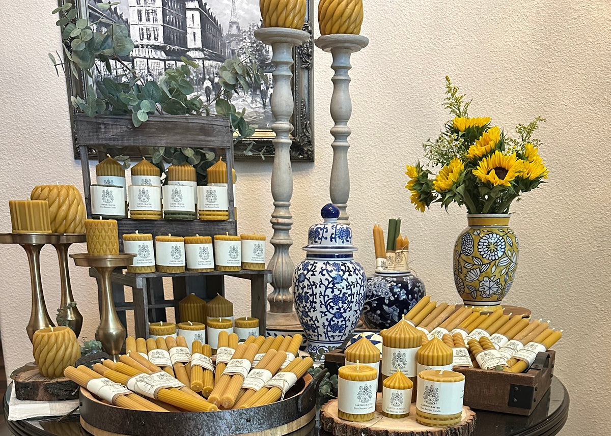 A styled display of artisan candles and wax products arranged on a round black pedestal table, with additional items in baskets and on shelves in a home-like interior. The products include many small white candles in glass jars, yellow beeswax taper candles, wrapped wax sticks, and a blue-and-white ceramic jar.