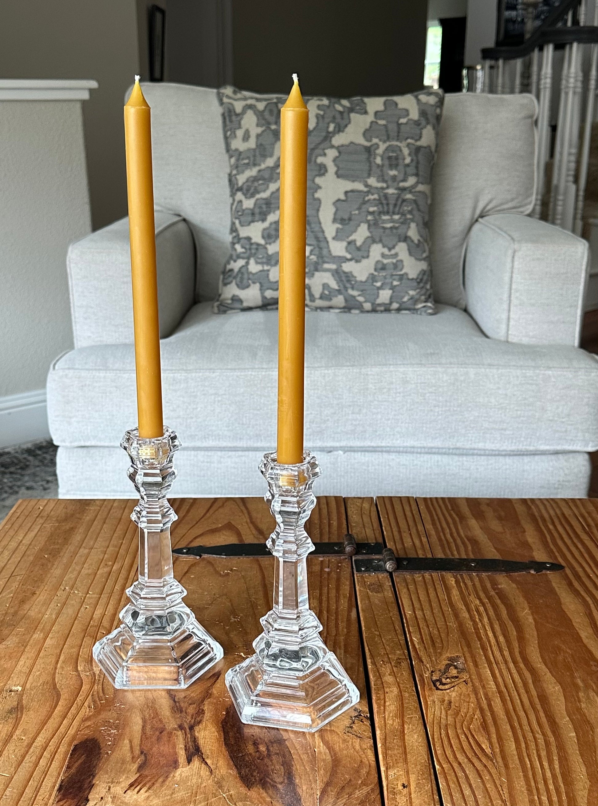 Two clear glass candlesticks with Modern Taper beeswax candles in Natural gold on a rustic wooden table in front of a gray chair.